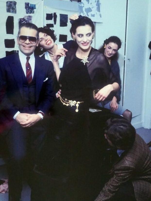 Karl Lagerfeld in schwarzer Kleidung zeigt auf Fotos an einer Wand mit Kunstwerken.